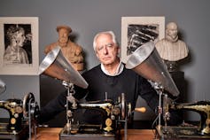 A portrait of a man with greying hair surrounded by statues, artworks and gramophone-like machines.