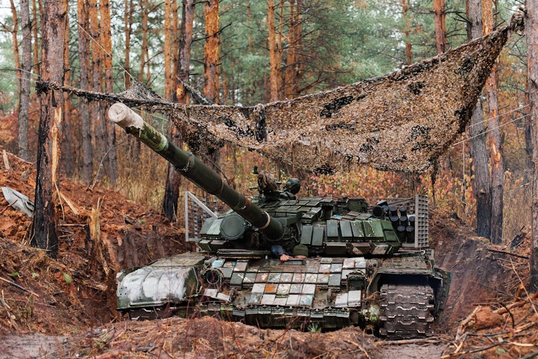 A tank with camouflage covering and surrounded by trees in the Donbas, Ukraine.