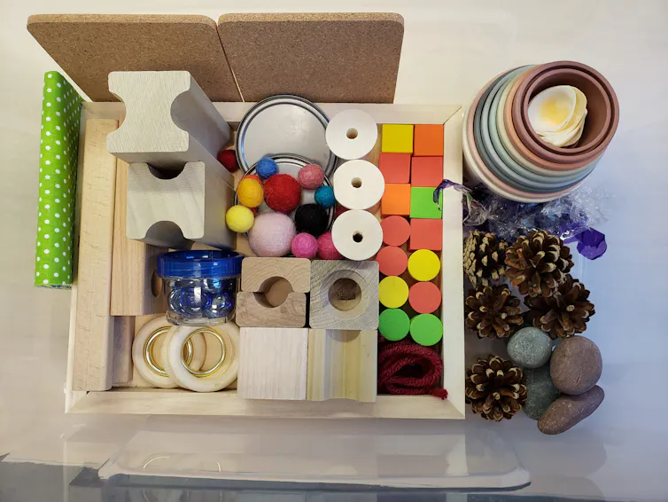 A cardboard box holding items like blocks, felt balls, pinecones, fabric.