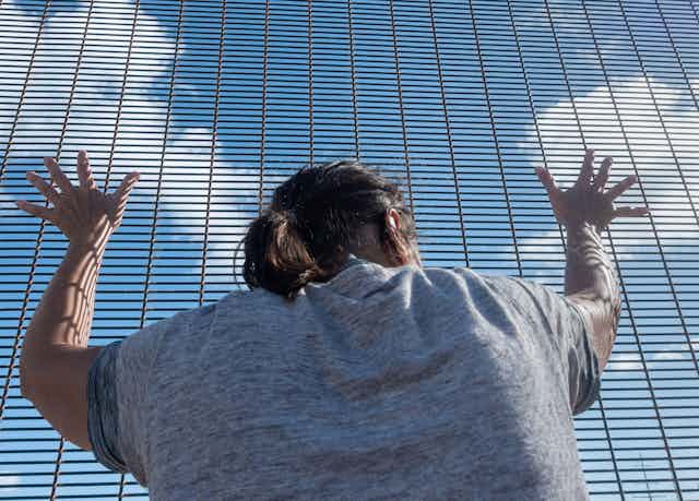 A woman clutches a metal fence and looks out to the blue skies beyond. 
