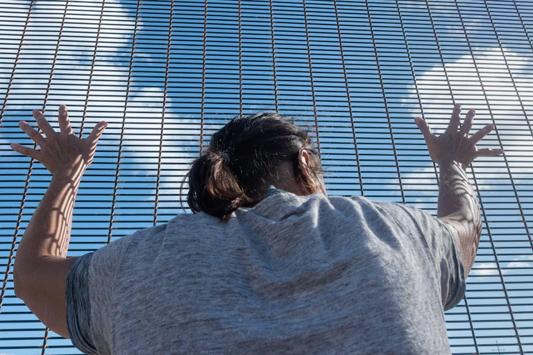 A woman clutches a metal fence and looks out to the blue skies beyond.