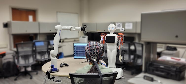 Person sitting in chair wearing electrode cap with a computer screen and robotic arms on a table in front of them