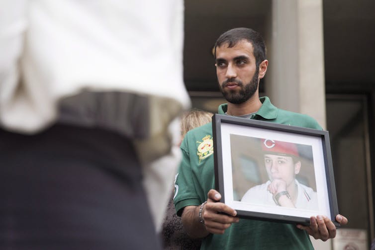 A man with a beard holds up a framed photo of another man.