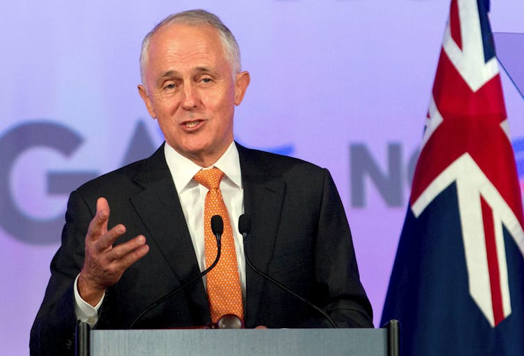 A grey-haired man in a dark suit and orange tie speaks into a microphone.