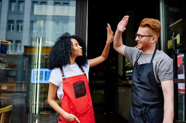 A man and a woman, who are both wearing aprons, high five in front of the glass doors of a store