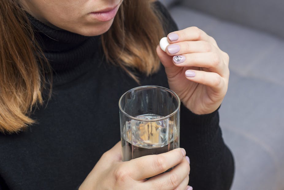 A young woman holding a glass of water in her right hand prepares to take a round, white pill.