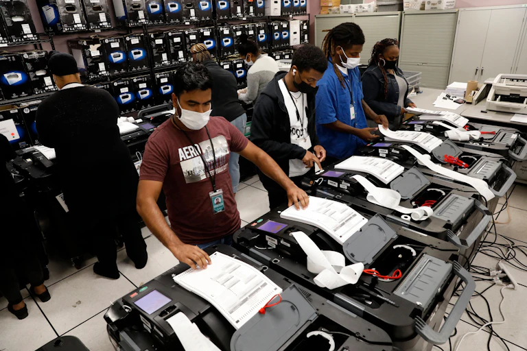 People wearing face masks stand in front of black machines that look like printers and feed paper into it.