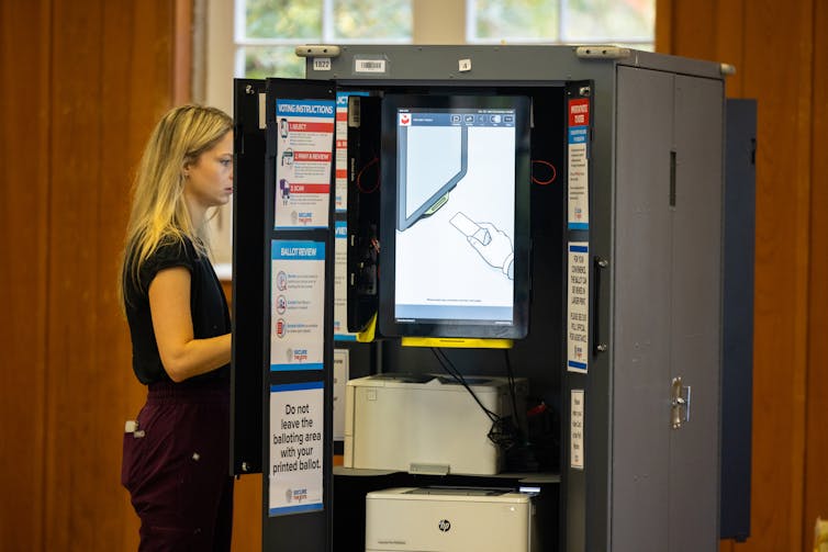 A blonde white woman stands facing an electronic voting booth.