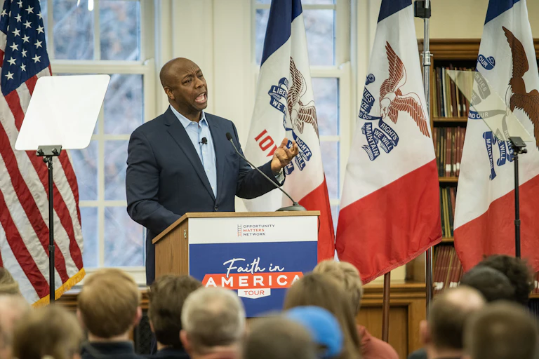 A man, with his left hand held chest high, open and facing up, stands behind a Faith in America tour lectern as he speaks to a seated group of people.