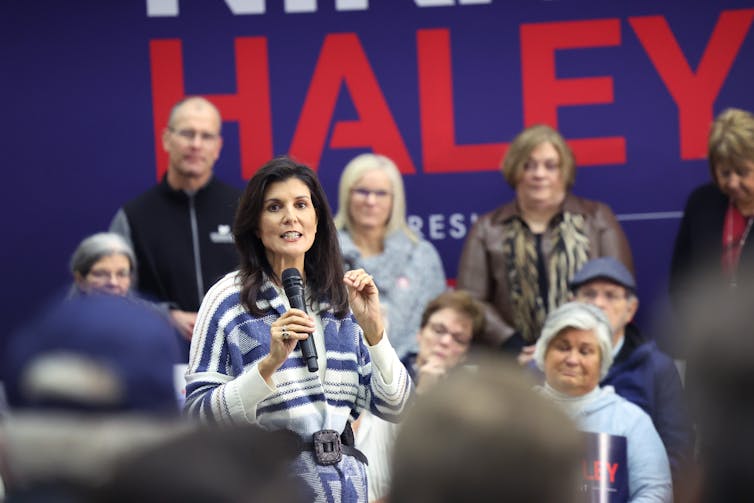 A standing woman, speaks into a microphone in her right hand, while she holds her left hand shoulder high with her fingers and thumbs in a pinched position. People sit in front of her and behind her. In the background is a purple sign with the name Haley