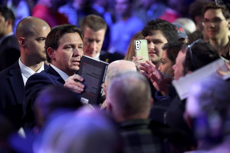 A suited man, surrounded by people, stands with his right arm outstretched and a book in-hand.