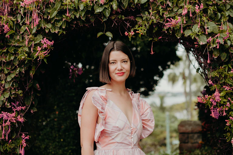 a woman in a pink dress stands under a trellis of pink flowers, subtly smiling