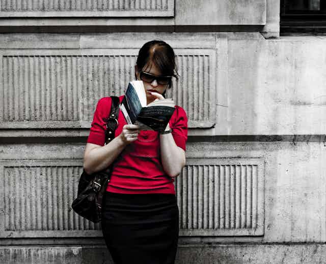 A woman reads a book leaning against a wall.