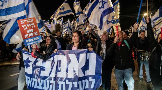 People at a protest at night wave Israeli flags and carry signs with Hebrew words.