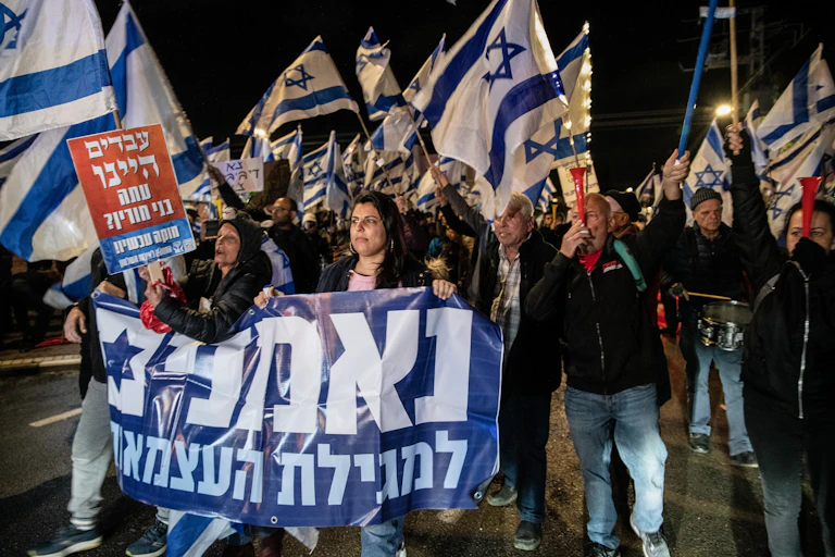 People at a protest at night wave Israeli flags and carry signs with Hebrew words.