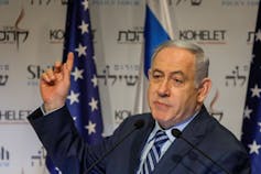 A gray-haired man stands next to the U.S. and Israeli flags while speaking at an event.