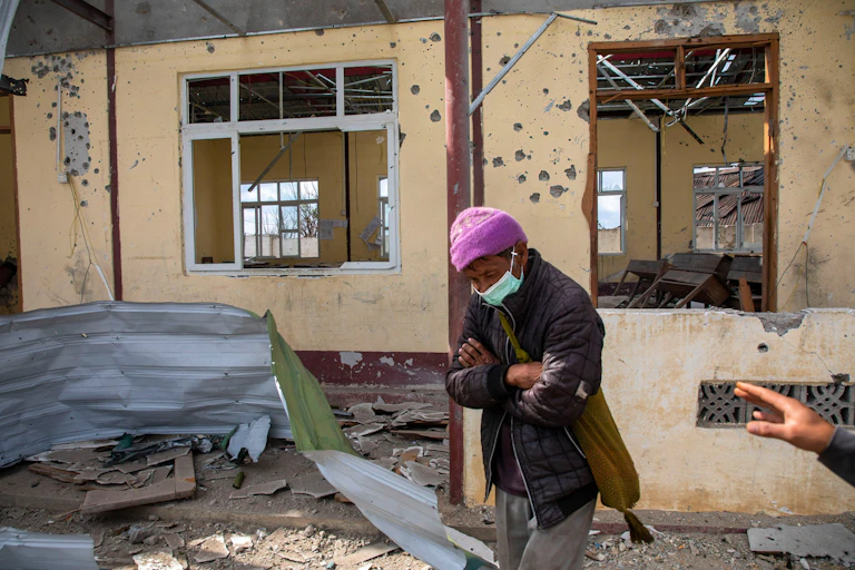 A man in a pink hat stands in front of a b uilding pockmarked by gunfire