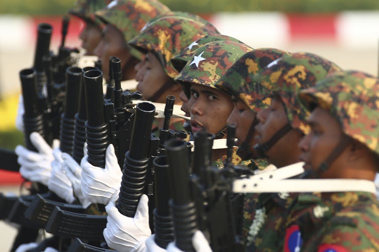 A line of men with camouflage helmets and guns. One looks at the camera.