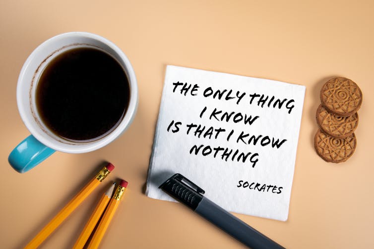 A coffee mug, pencils, pen and cookies next to a note reading 'The only thing I know is that I know nothing – Socrates.'
