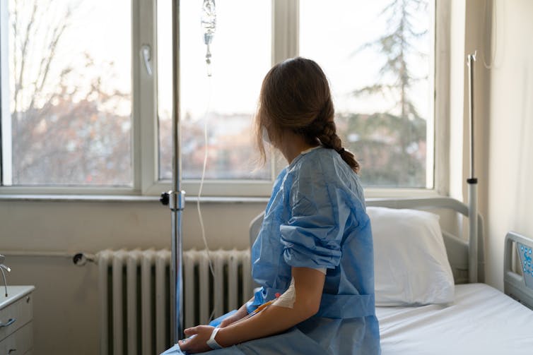Patient in hospital gown with IV sitting on bed, looking out window