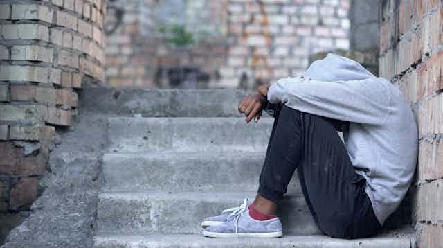 Dressed in dark sweats, sneakers, and with a gray hoodie covering his face, a forlorn young Black man sits on the steps outside his apartment building.