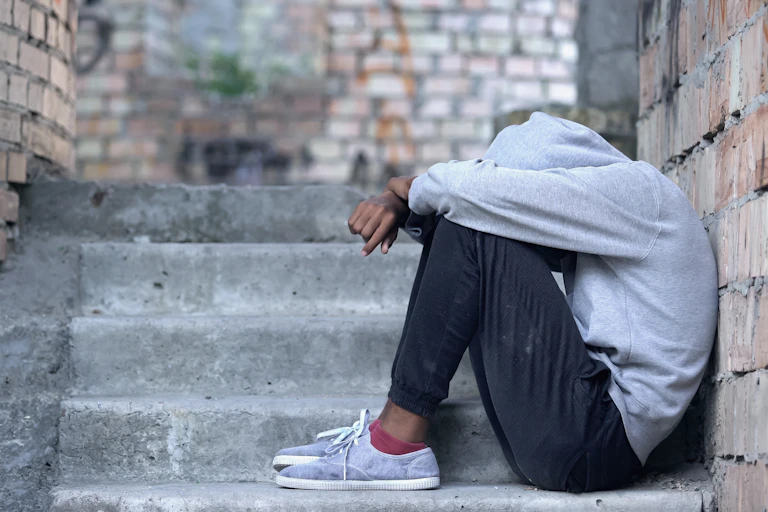 Dressed in dark sweats, sneakers, and with a gray hoodie covering his face, a forlorn young Black man sits on the steps outside his apartment building.