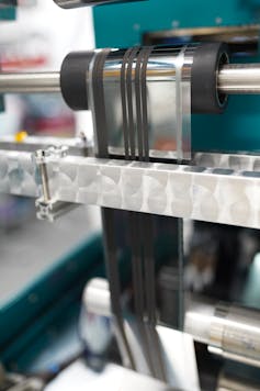 A roll of thin silver film passes through a machine on reels.