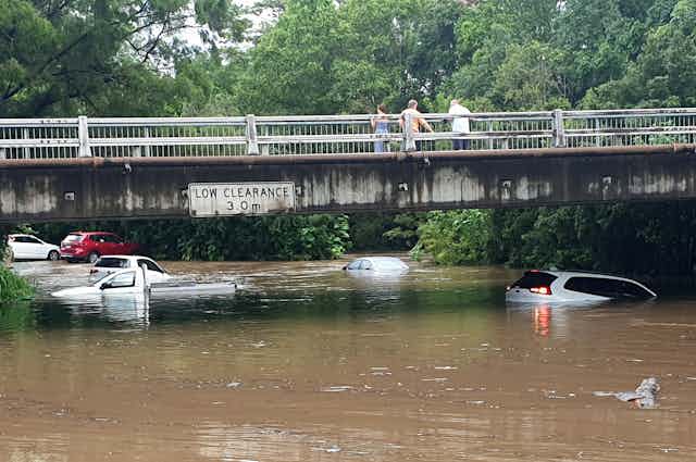 Cars submerged beneath floodwaters under a bridge with pedestrians looking down from above.