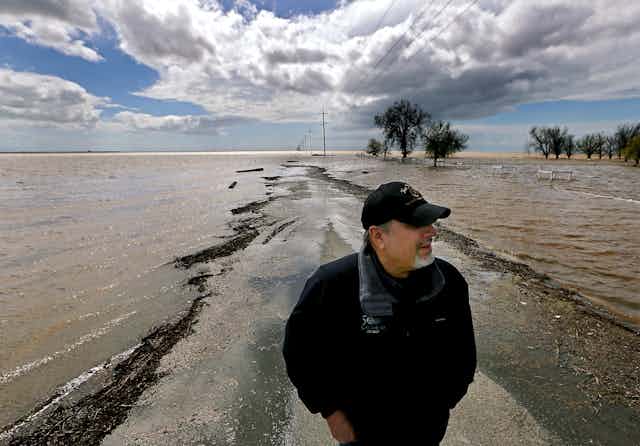 A man stands on a dry spot of a farm road with water on both sides and flooded fields.