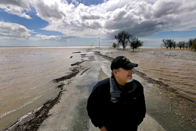 A man stands on a dry spot of a farm road with water on both sides and flooded fields.