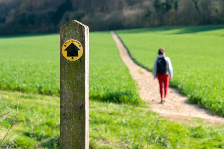 A public footpath post in the foreground and a hiker travelling a long path into the distance.