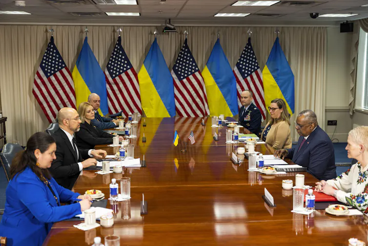 US and Ukrainian officials sit around a boardroom table in a room decorated by US and Ukraine flags.