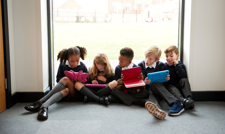 Five young children in school uniforms are sitting together, looking at the screens of their tablets