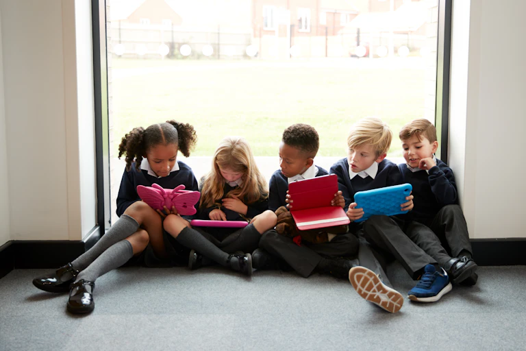 Five young children in school uniforms are sitting together, looking at the screens of their tablets