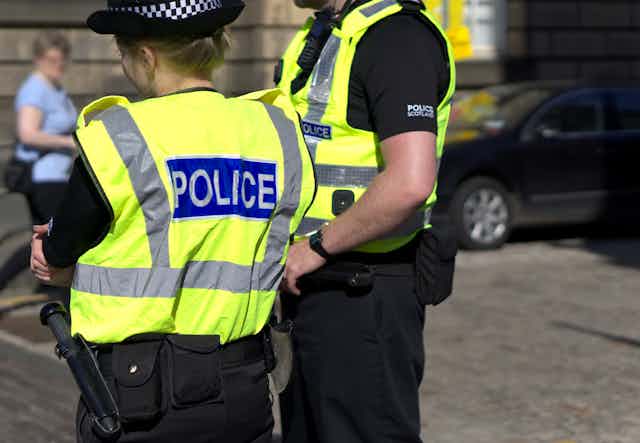 One female and one male police officer standing on a city street, viewed from behind, wearing hi vis vest that reads POLICE.