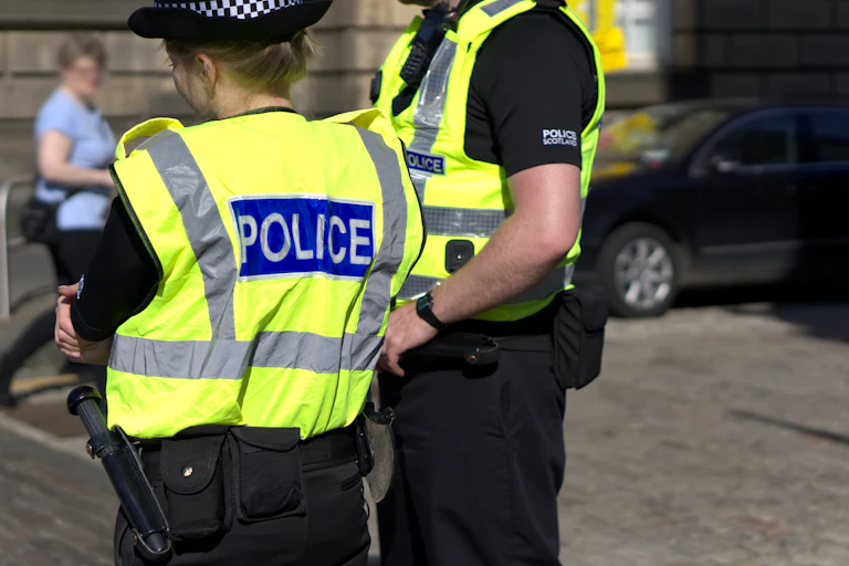 One female and one male police officer standing on a city street, viewed from behind, wearing hi vis vest that reads POLICE.