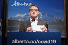 A man in glasses holds up a mask. The podium in front of him reads alberta.ca/covid19.