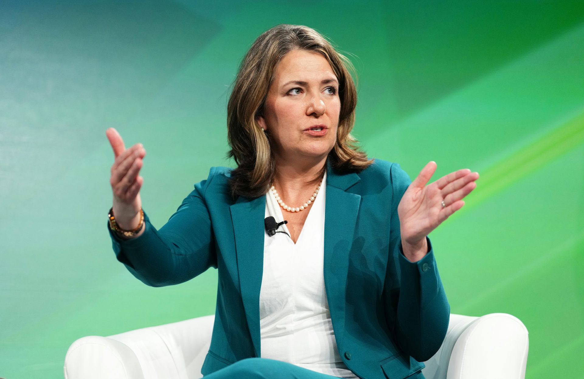 A dark-haired woman in a green suit and white shirt gestures as she speaks while sitting in a white easy chair.