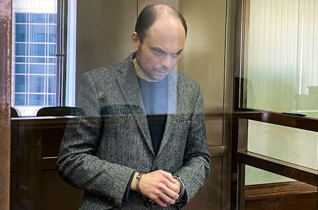 A balding man in a grey jacket stands with his hands clasped and his head down