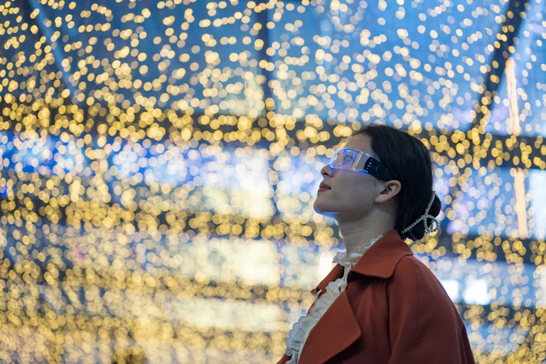 A woman with dark hair and safety goggles in an orange coat looks up toward a clear ceiling dotted with tiny lights.