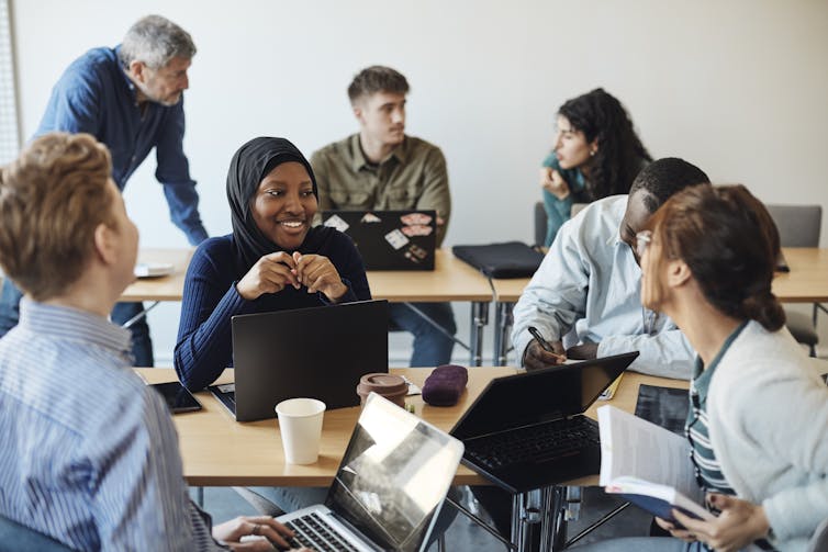 Half a dozen students, including one in a hijab, chat at long tables, with a professor at the back of the photo.