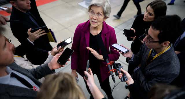 A woman in black pants and a fuschia jacket with short gray hair speaks to a gaggle of reporters.