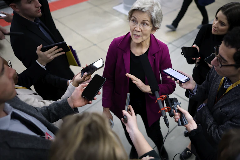 A woman in black pants and a fuschia jacket with short gray hair speaks to a gaggle of reporters.