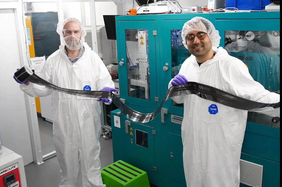 Two men wearing white overalls and hair nets hold up a long piece of black film