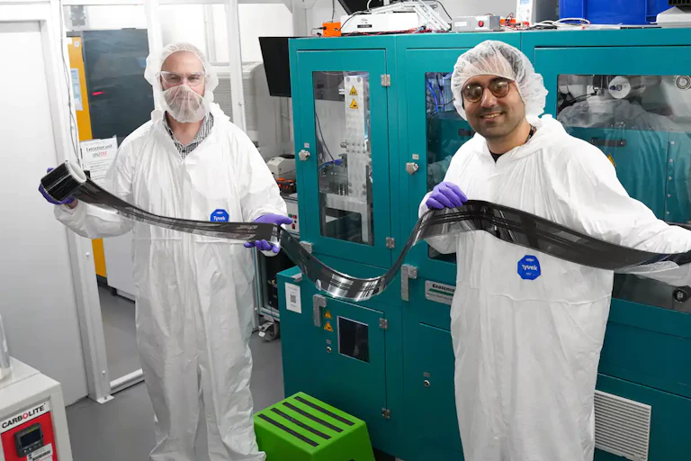 Two men wearing white overalls and hair nets hold up a long piece of black film