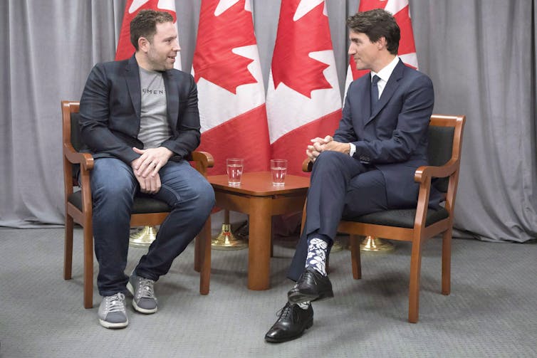 Two men, one wearing a suit and one in casual wear, chat with one another while seated. Canadian flags stand in the background.