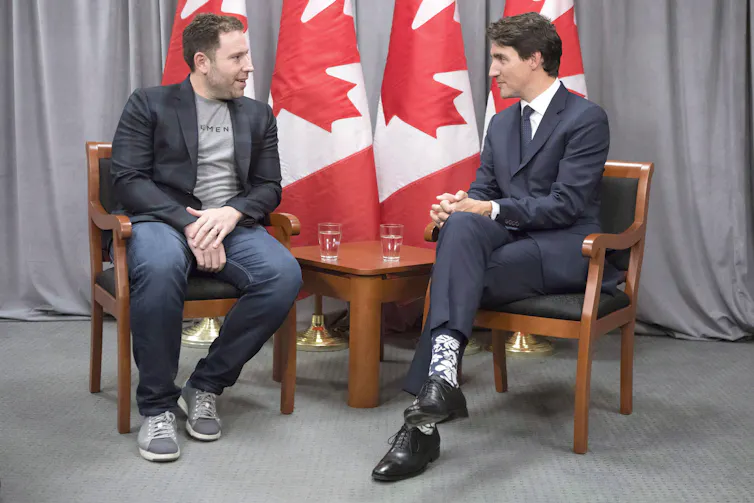 Deux hommes, l’un portant un veston et l’autre une tenue décontractée, sont assis et discutent.Des drapeaux canadiens apparaissent en arrière-plan