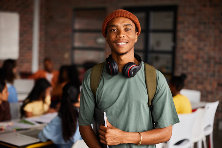 A college-age man with headphones around his neck and an army-green backpack looks at the viewer as he clutches a laptop.