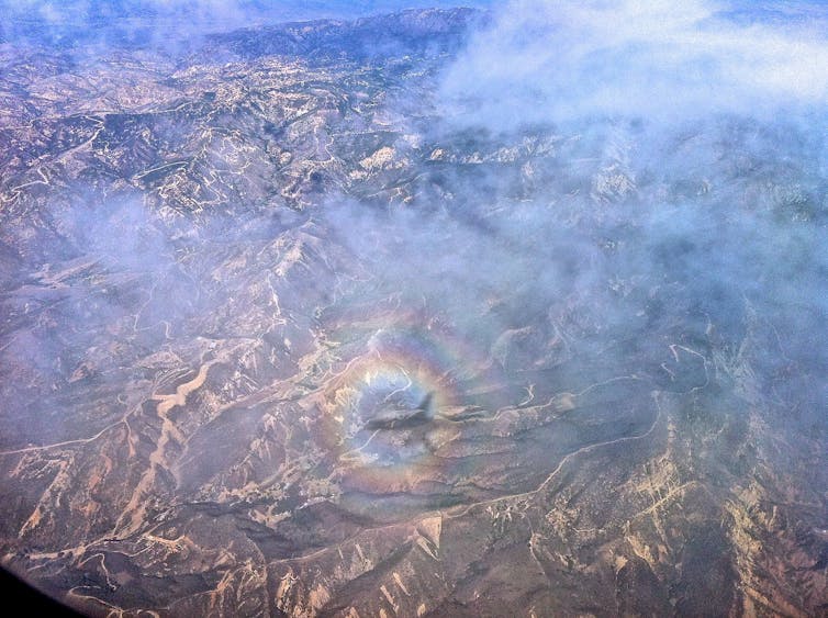An airplane's shadow has a circular rainbow around it as it flies over mountains.