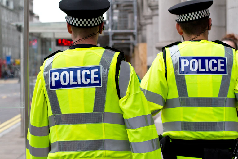 The backs of two police officers wearing high visibility yellow jackets. They are wearing police headgear and have wires in their right ears.